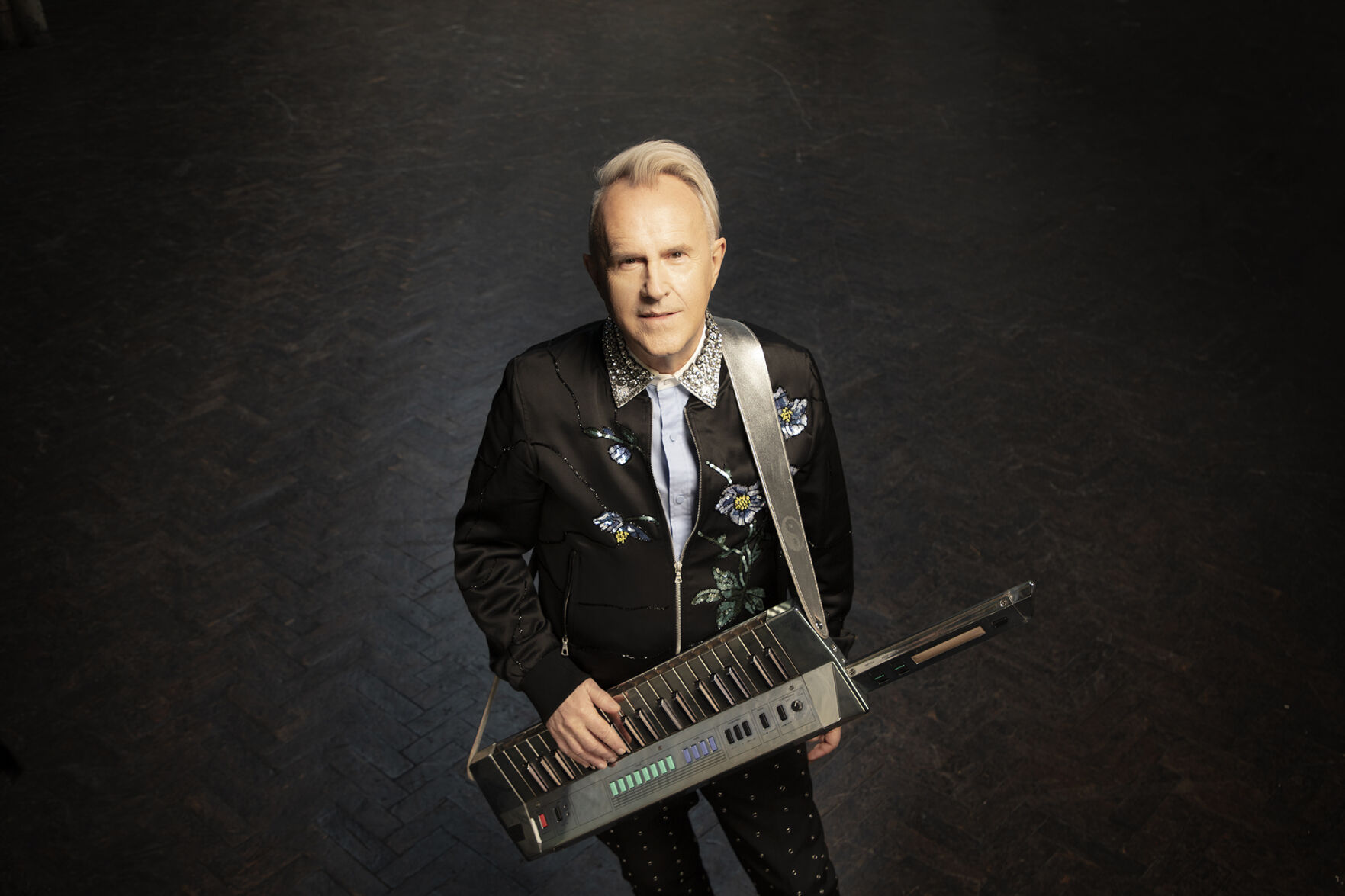 Howard Jones press pic artist wears a dark color Western shirt holds a keytar and smiles at the camera